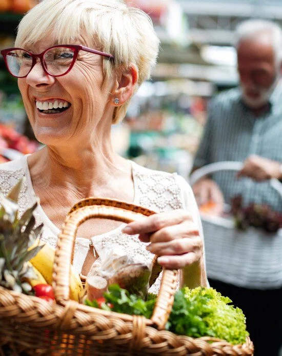 Woman smiles at market with basket full of fresh produce and another shopper nearby.
