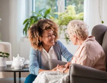 Female home caregiver talking with senior woman, sitting in living room and listening to her carefully. caregiver smiles while helping senior woman who sits in a reclining chair