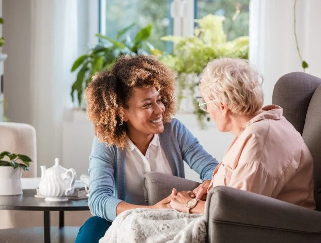 caregiver smiles while helping senior woman who sits in a reclining chair