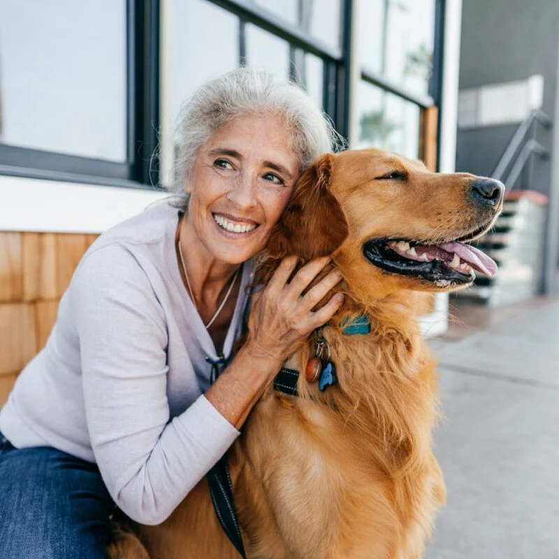 senior woman smiles and kneels down to pet her smiling dog