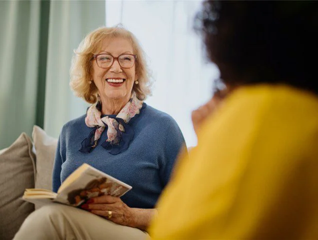 senior woman smiles while reading book and conversing with friend