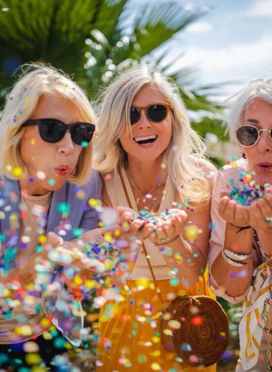 Three women blow colorful confetti outdoors on sunny day.