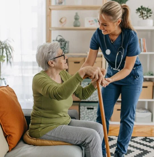 caregiver smiles and assists senior woman seated on a couch in her apartment