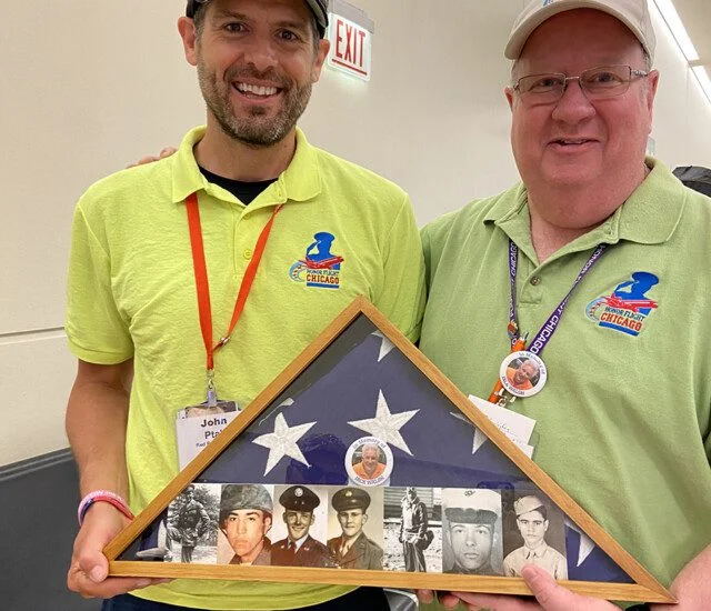 Two men in matching shirts hold a framed American flag with veteran photos.