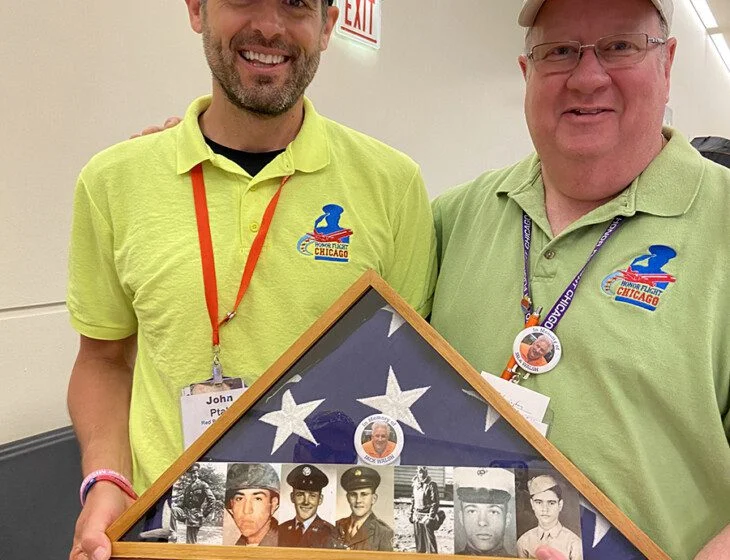 Two men in matching shirts hold a framed American flag with veteran photos.