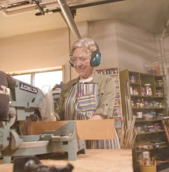 A smiling individual works with wood using power tools in a workshop setting.