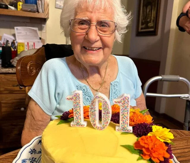 Elderly woman smiling with a 101 cake, adorned with colorful flowers, in a kitchen setting.