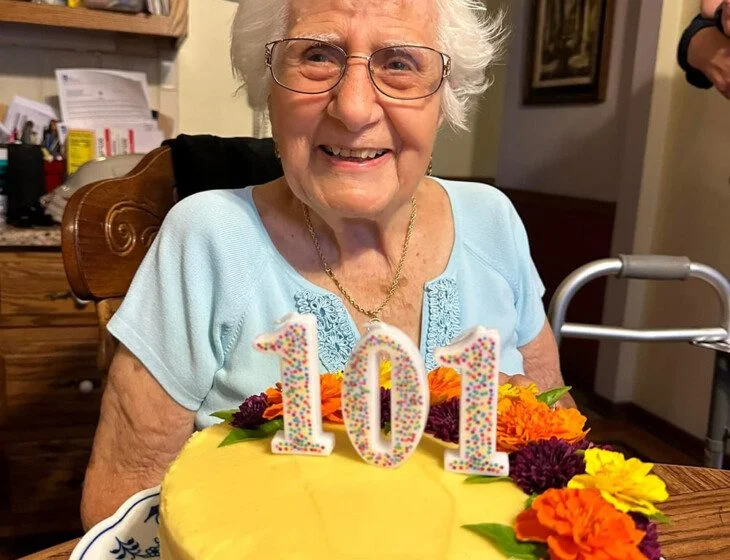 Elderly woman smiling with a 101 cake, adorned with colorful flowers, in a kitchen setting.