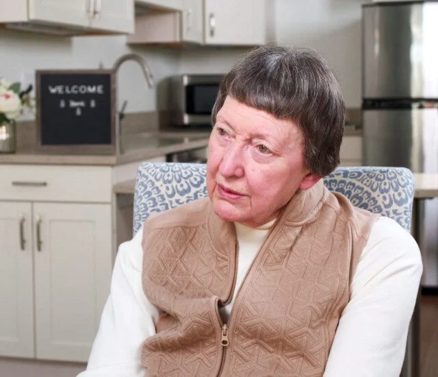 Woman in a beige vest sitting in a kitchen, looking thoughtfully.