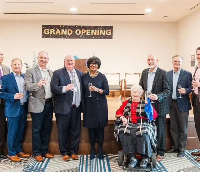 Group celebrating a grand opening, holding glasses, banner in background.