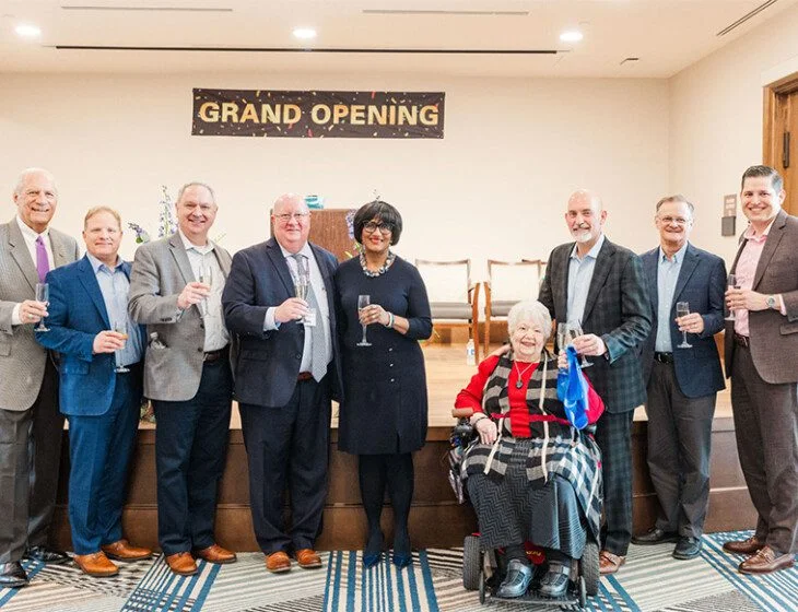 Group celebrating a grand opening, holding glasses, banner in background.