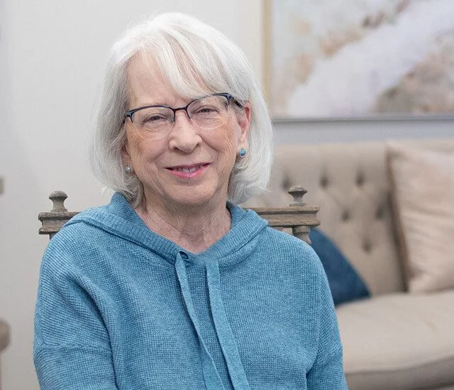 A smiling older woman with white hair and glasses wears a blue sweater indoors near furniture.