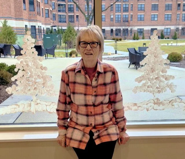 Elderly woman in plaid shirt, sitting by a window with decorative tree designs.