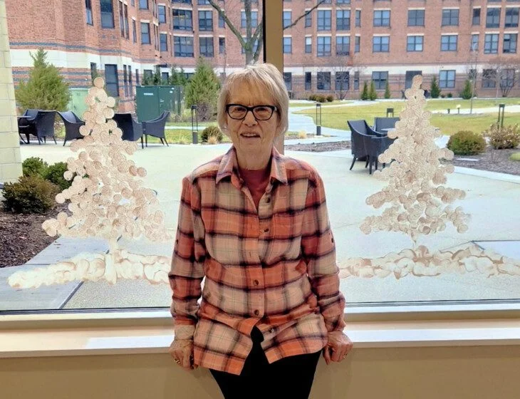 Elderly woman in plaid shirt, sitting by a window with decorative tree designs.