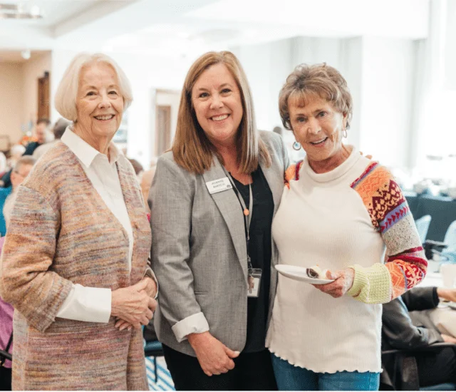 Three women smiling at a luncheon event indoors.