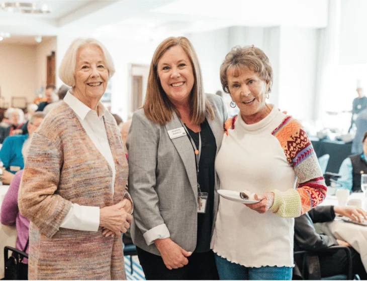 Three women smiling at a luncheon event indoors.