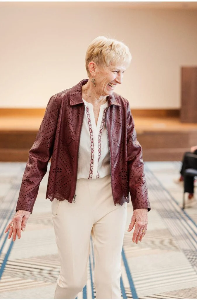 Smiling woman in burgundy jacket and white pants walking indoors.