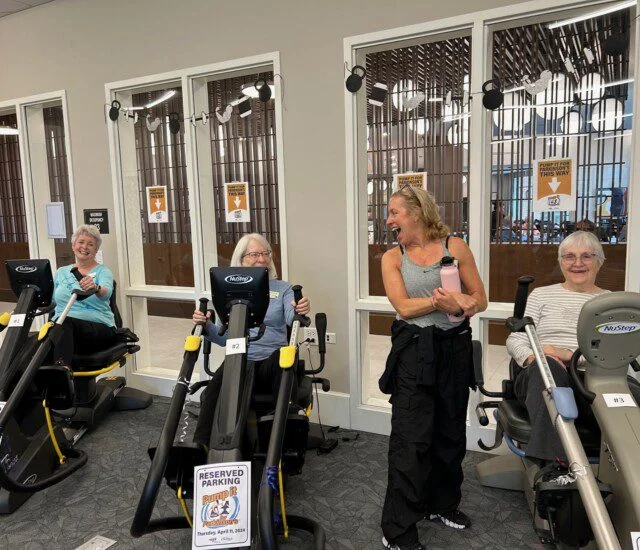 Three women on exercise machines, smiling, with instructor nearby in a gym setting.