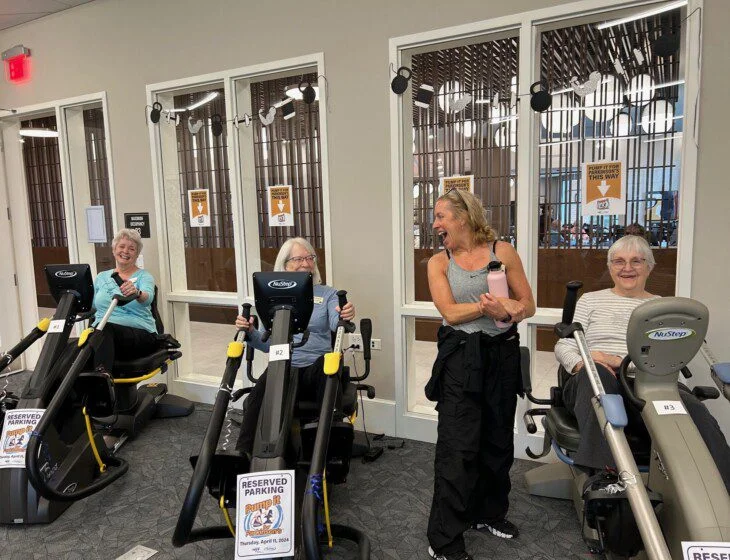 Three women on exercise machines, smiling, with instructor nearby in a gym setting.