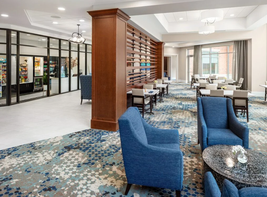 Modern lobby with blue chairs, tables, and patterned carpet, adjacent to a glass-walled shop.