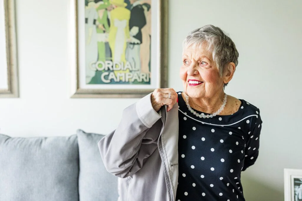 Smiling woman in a polka dot dress holding a coat, with vintage poster in background.