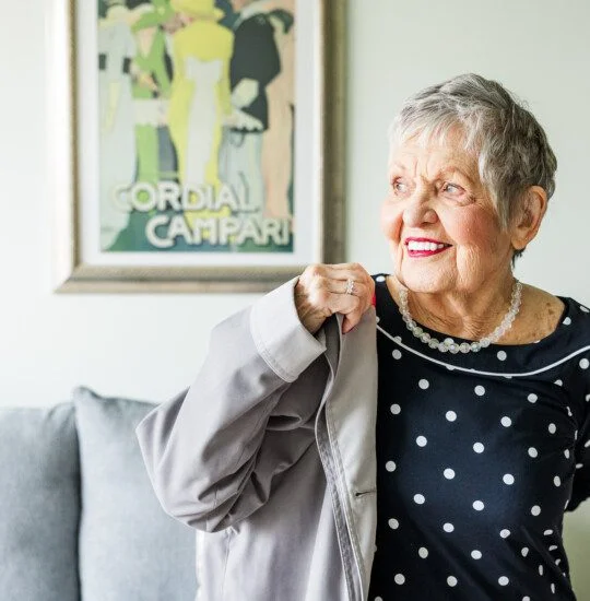 Smiling woman in a polka dot dress holding a coat, with vintage poster in background.