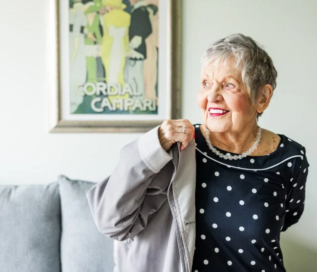 Smiling woman in a polka dot dress holding a coat, with vintage poster in background.