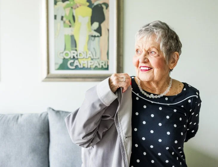 Smiling woman in a polka dot dress holding a coat, with vintage poster in background.