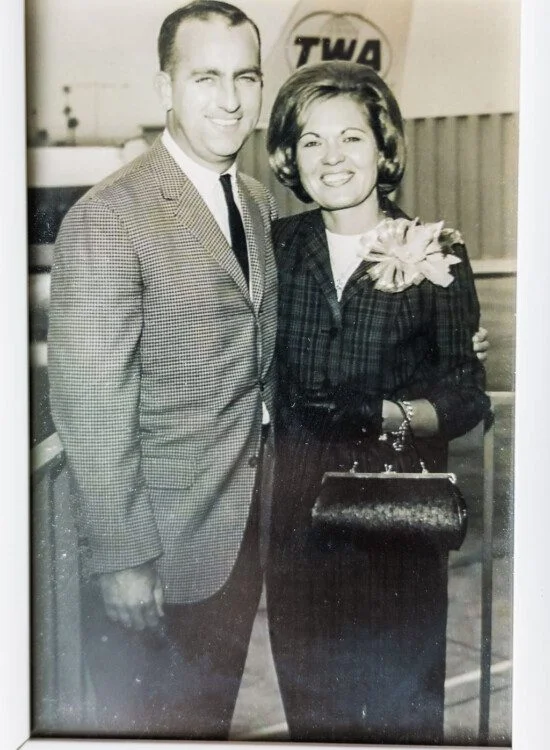 Smiling couple in vintage attire at an airport with TWA logo in the background.