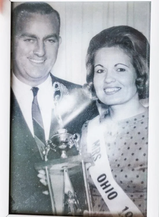Man and woman smiling, holding a trophy. Woman wearing Mrs. Ohio sash. Black and white photo.