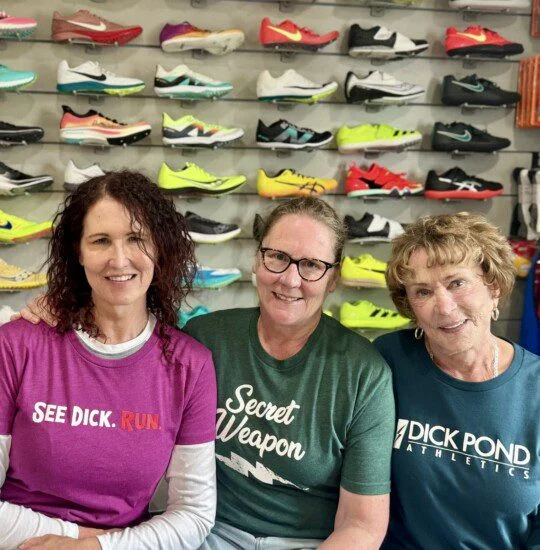Three women smiling in front of a wall display of colorful athletic shoes.