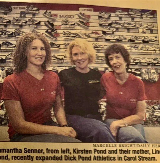 Three women sit in front of a wall of shoes at a sports store.