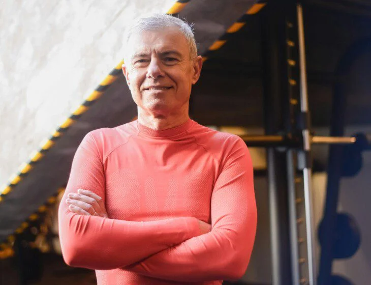 Man in red workout shirt, smiling, arms crossed in a gym.