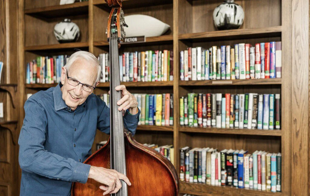 Elderly man playing a double bass in front of a bookshelf in a library.