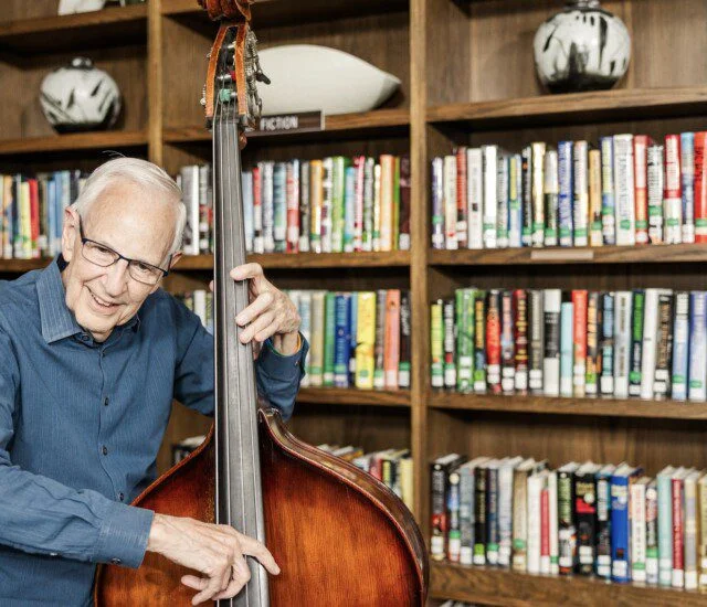 Elderly man playing a double bass in front of a bookshelf in a library.
