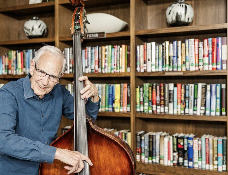 Elderly man playing a double bass in front of a bookshelf in a library.