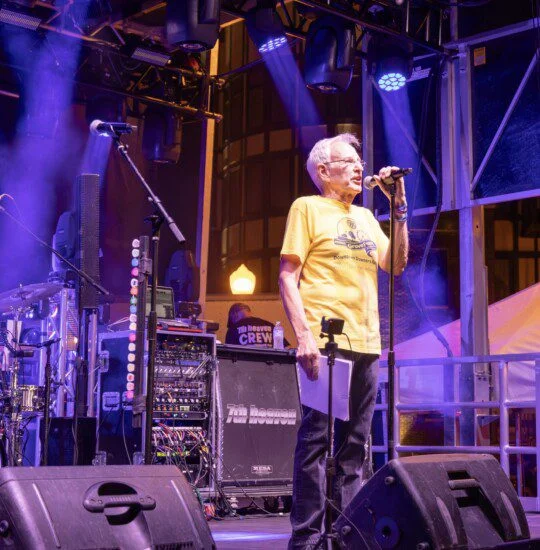 Man in yellow shirt speaking on stage at an event with music equipment.