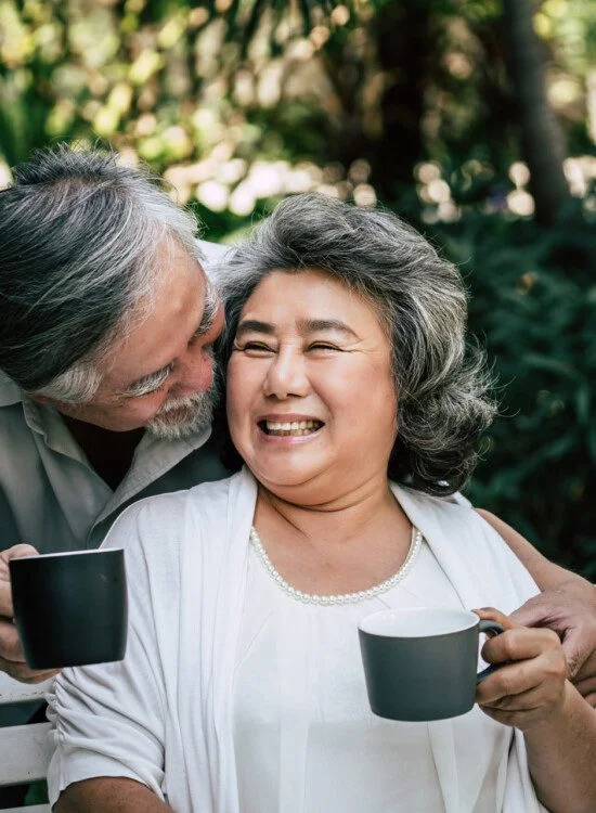 Elderly couple smiling and holding mugs in a garden setting.