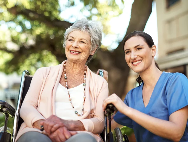 Elderly woman in wheelchair with smiling caregiver outdoors.