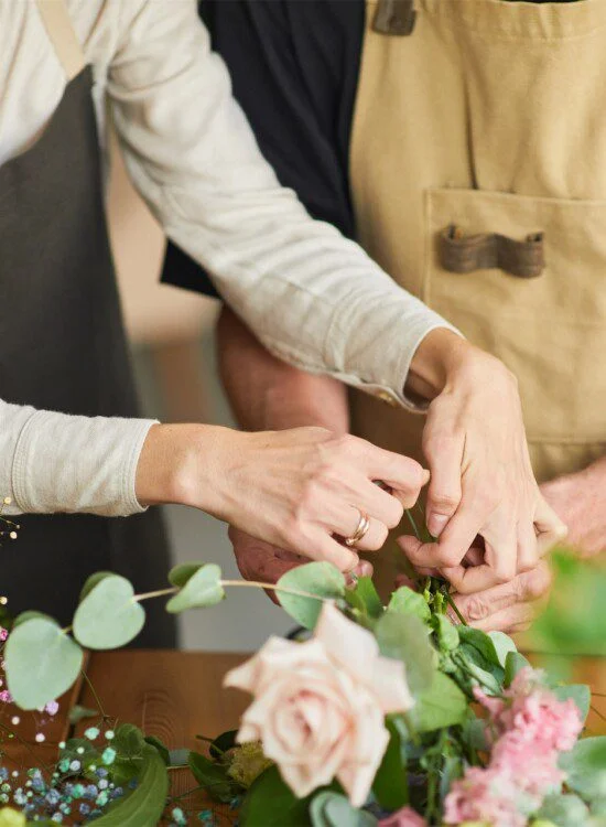 Hands arranging flowers with eucalyptus leaves on a table.