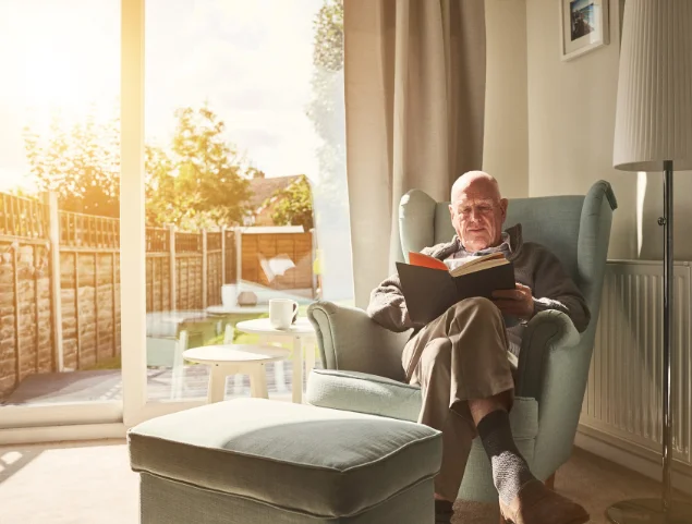 Elderly man reading by sunny window in cozy living room.