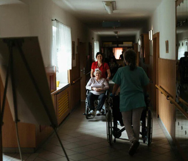 Nurses assist elderly woman on wheelchair through hallway.