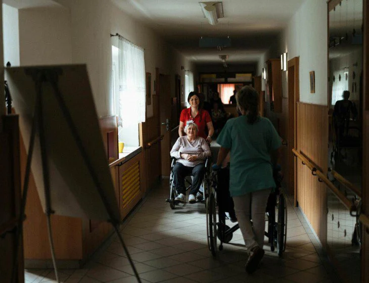 Nurses assist elderly woman on wheelchair through hallway.