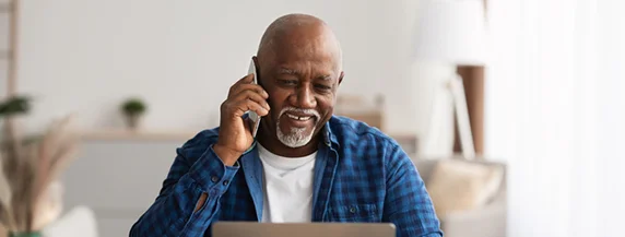 Mature Man Talking On Cellphone And Using Laptop Man in blue shirt smiling and talking on phone while using a laptop at home.