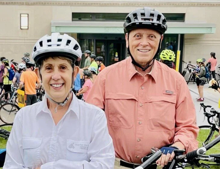Two people in helmets smiling, holding bikes at an outdoor event with others in the background.