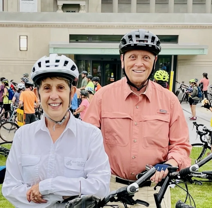 Two people in helmets smiling, holding bikes at an outdoor event with others in the background.