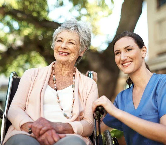 Elderly woman in wheelchair with smiling caregiver outdoors.