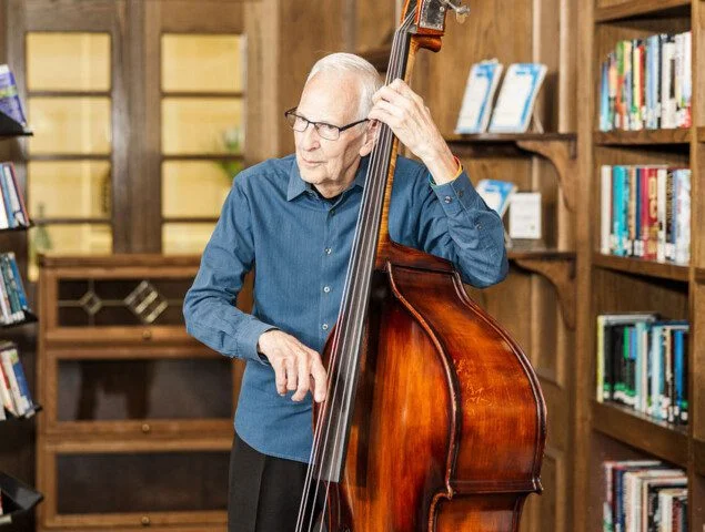 Elderly man playing double bass in a library setting.