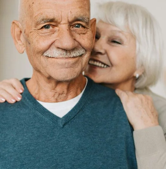 Elderly couple smiling, the woman embracing the man from behind.