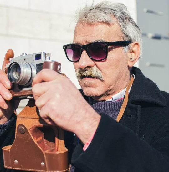 Older man with sunglasses photographing outdoors with a vintage camera.